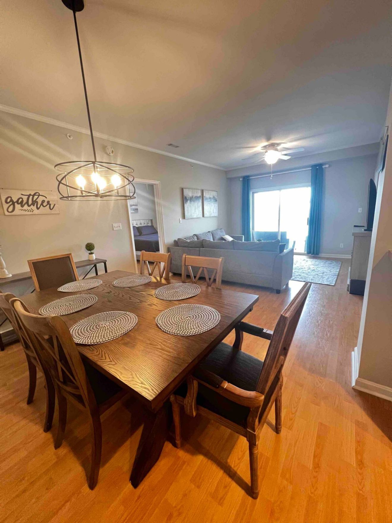 A dining area with a wooden table and chairs, a ceiling fan, and a wall with a 'gather' sign.