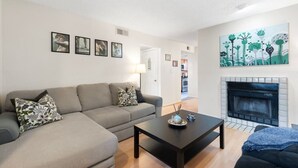 A living room with a sofa, a coffee table, and a fireplace. There are framed pictures on the wall and a kitchen in the background.