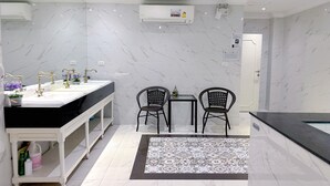 A modern bathroom with a marble backsplash, a white vanity with gold faucets, two black wicker chairs, and a patterned rug.