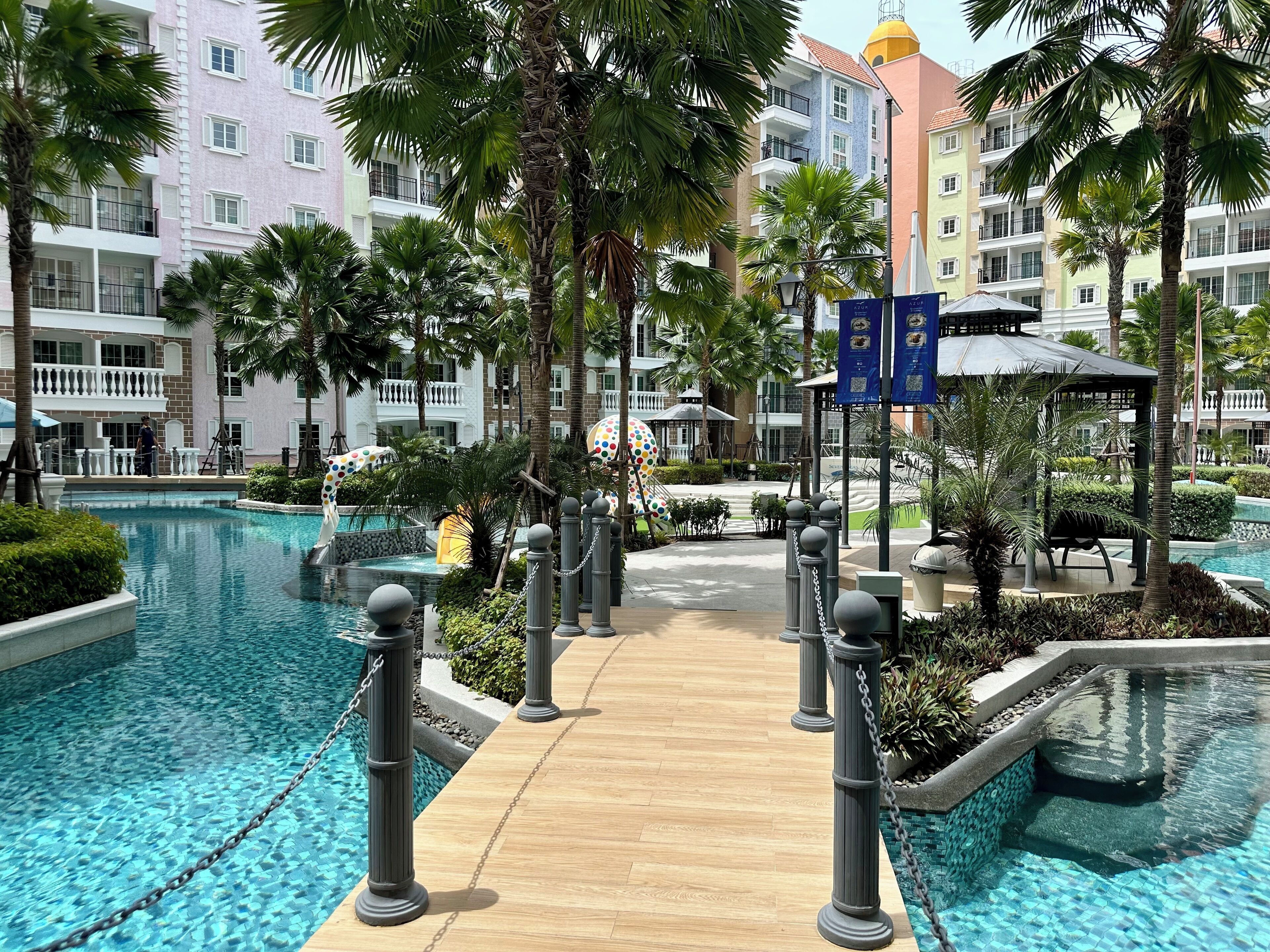 Zona de piscina en un resort con terraza de madera, palmeras y un edificio de varios pisos con balcones.