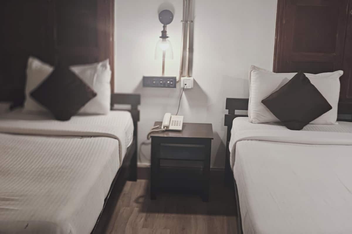 Ein Hotelzimmer mit zwei Einzelbetten, jeweils mit Kissen und Nachttisch. An der Wand hängt eine Lampe und auf dem Tisch steht ein Telefon.