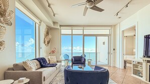 Ein geräumiges Wohnzimmer mit großem Fenster, Deckenventilator, einer Couch, einem Couchtisch und einem Fernseher.