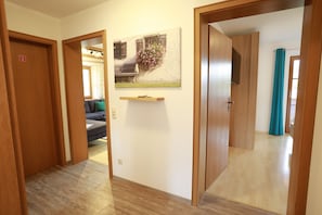 A hallway with wooden doors, a painting of a house with flowers, and a glimpse of a living room with a sofa and cushions.