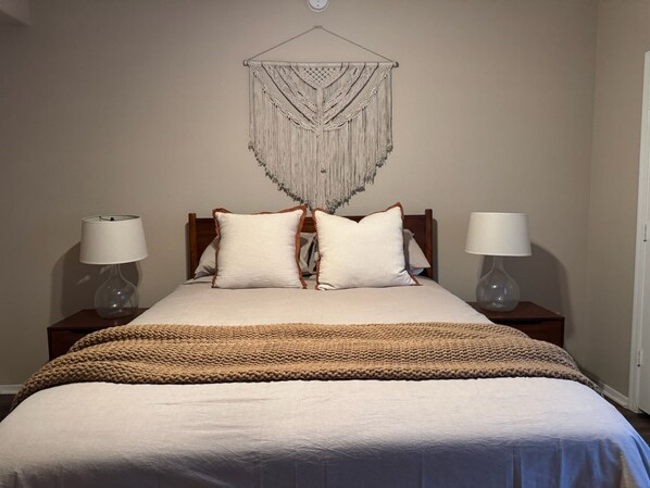Ein ordentlich bezogenes Bett mit einer weißen und beigen Decke, zwei Nachttischlampen und einem Wandbehang aus Makramee.