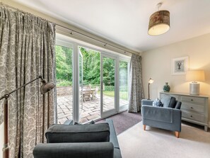 Une pièce comprenant un canapé, une lampe, une table d’appoint avec un vase, un tableau au mur et, par une porte vitrée, une vue sur un espace extérieur aménagé avec une table et des chaises.