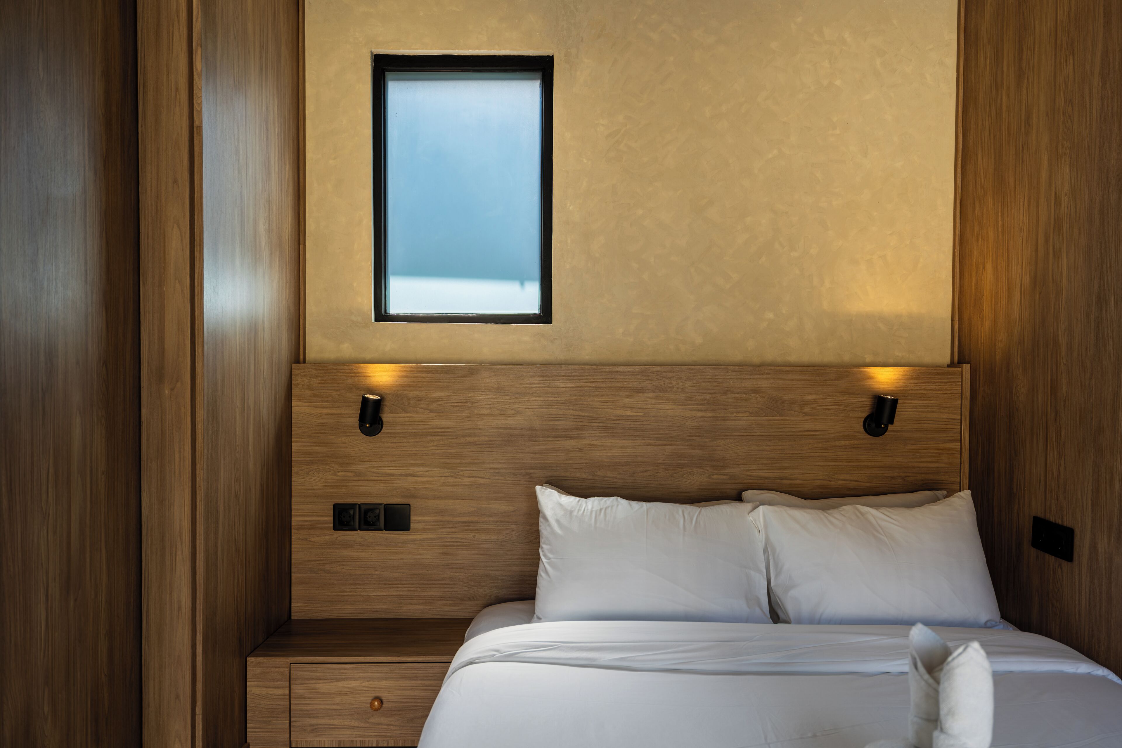 A wooden-framed bed with white bedding, a nightstand, and two wall-mounted reading lights.