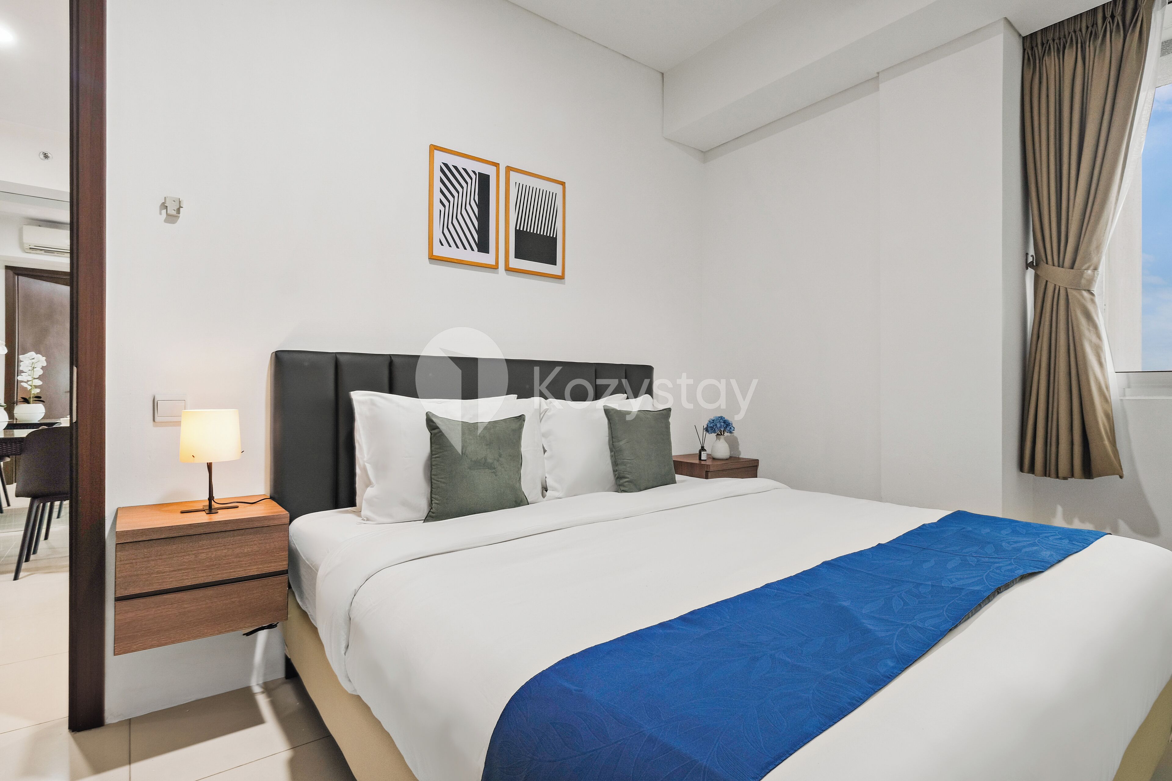 A modern hotel room with a neatly made bed, a bedside table with a lamp, and two framed pictures on the wall.