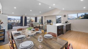 A spacious dining area with a wooden table set for six, a kitchen with a modern design, and large windows offering views of the outdoors. - Mountain Views on Sainsbury (Queenstown)