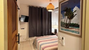 A hotel room with a bed, a TV mounted on the wall, a painting of a beach scene, and a door with a wooden frame. - Charming 1-bedroom apartment in lovely Papeete entrance. (Unknown)