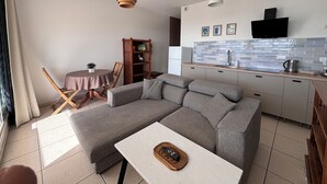 A modern living room with a grey sectional sofa, a small round table, and a white coffee table. There is a wooden bookshelf, a dining area with a table and chairs, and a kitchen with a tiled backsplash and a flat-screen TV. - Charming 1-bedroom apartment in lovely Papeete entrance. (Unknown)