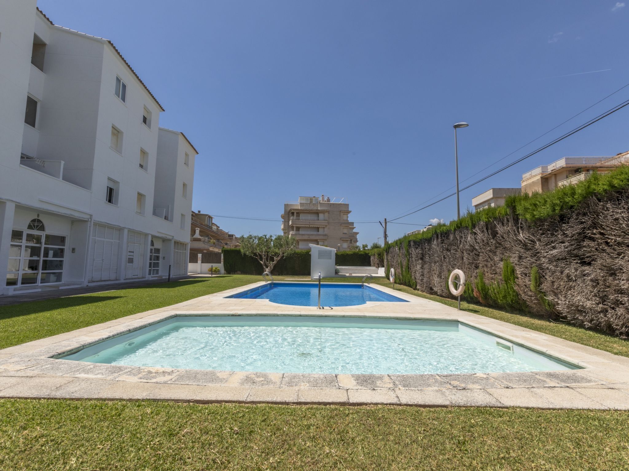 A swimming pool surrounded by a well-maintained lawn, with a building featuring multiple balconies and a clear blue sky in the background.