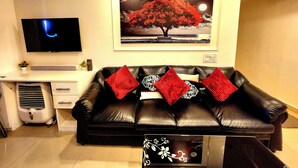 A living room with a black leather sofa, a flat-screen TV, a framed picture of a tree, and a small table with a floral design. - Xanadu Studio Apartment # 902 (Kolkata)