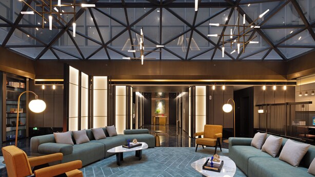 Eine moderne Lobby mit hoher Decke, einem geometrischen Deckendesign und einer zentralen Sitzecke mit Sofas und Sesseln.