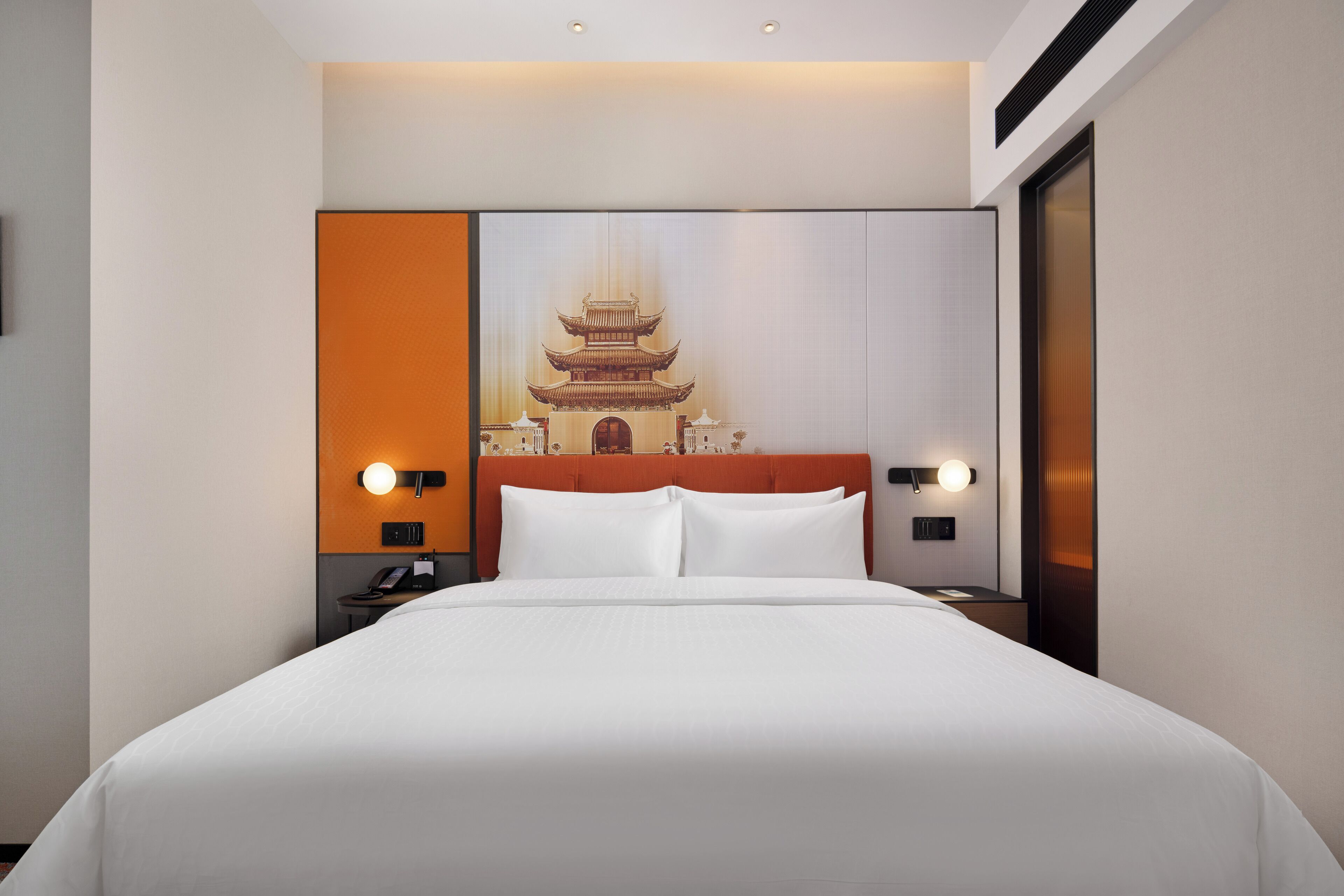 Una habitación de hotel con una cama grande, un cabecero con una pintura de una pagoda, dos mesitas de noche con lámparas y un aparador con un teléfono.