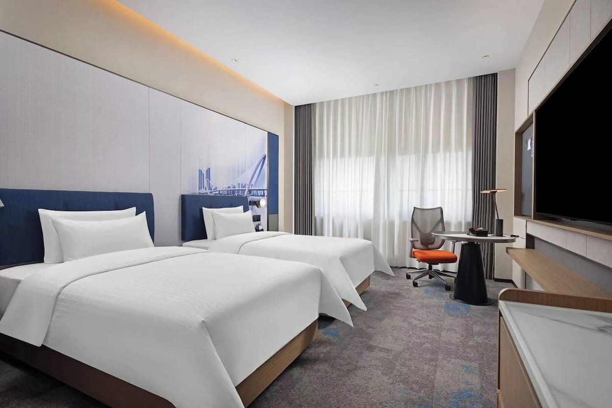 Une chambre d’hôtel avec deux lits, un bureau, une chaise, une télévision et une grande fenêtre avec des rideaux.