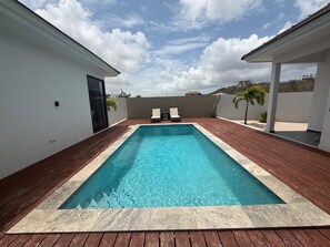 A rectangular swimming pool with a wooden deck surrounding it, two lounge chairs, and a view of a hilly landscape under a partly cloudy sky. - Villa Milamor Curacao - Harmonie, Grote Berg (Grote Berg)