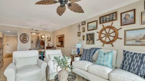 A living room with a white sofa, a coffee table with a vase, and a ceiling fan. - Sail Inn SaintSimons-ground unit,beach access,pool (St. Simons Island)