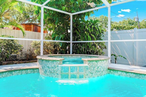 A swimming pool with a hot tub, surrounded by a white fence and lush greenery.