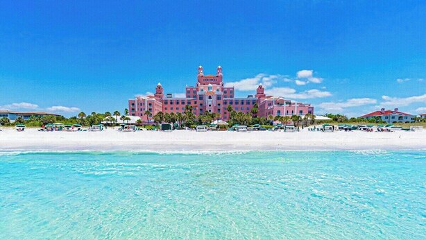 A large pink resort hotel with multiple buildings, a beach in the foreground, and clear turquoise water.