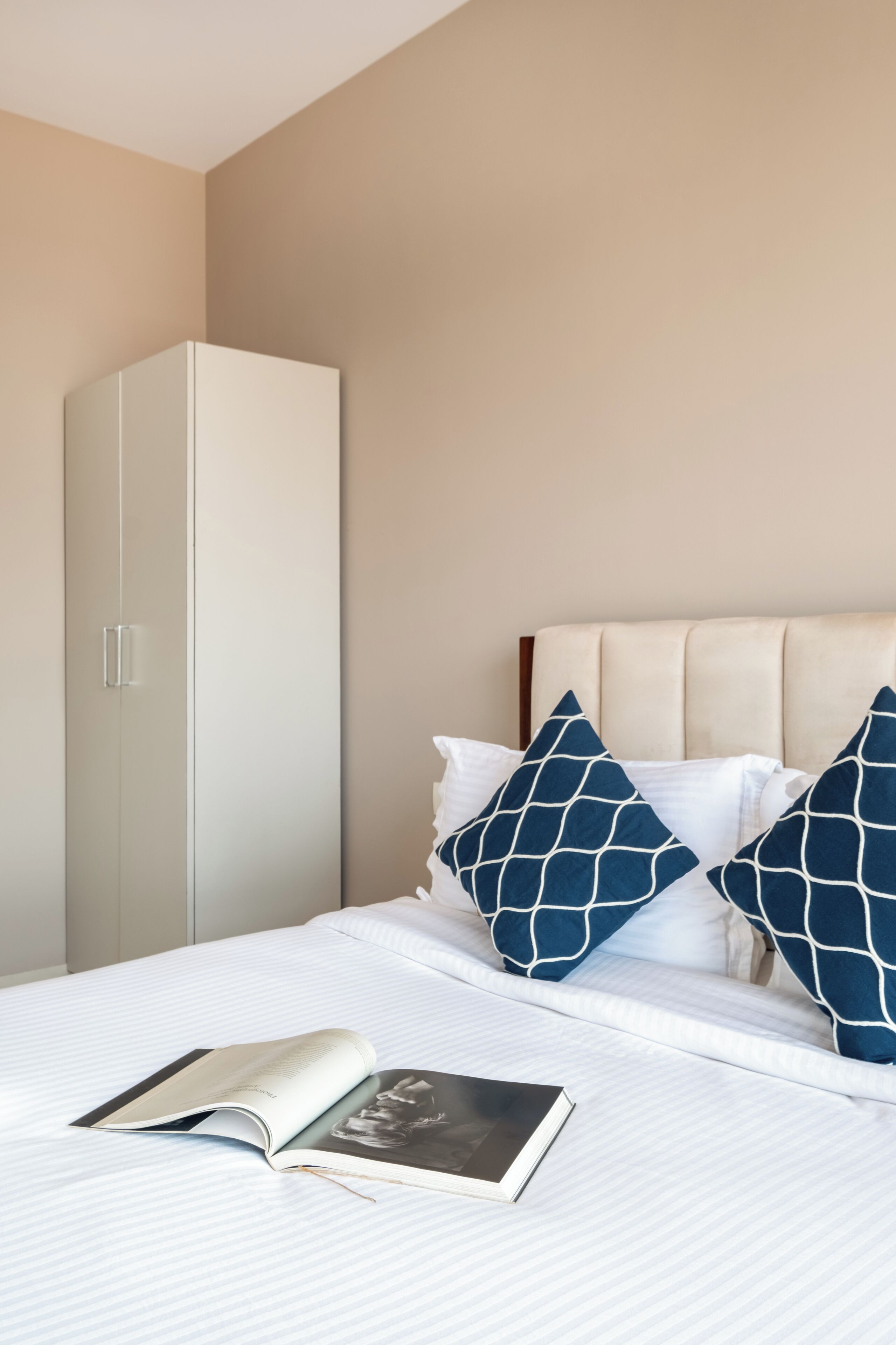 Ein ordentlich bezogenes Bett mit Buch und Kissen, ein weißer Kleiderschrank und eine beige Wand.