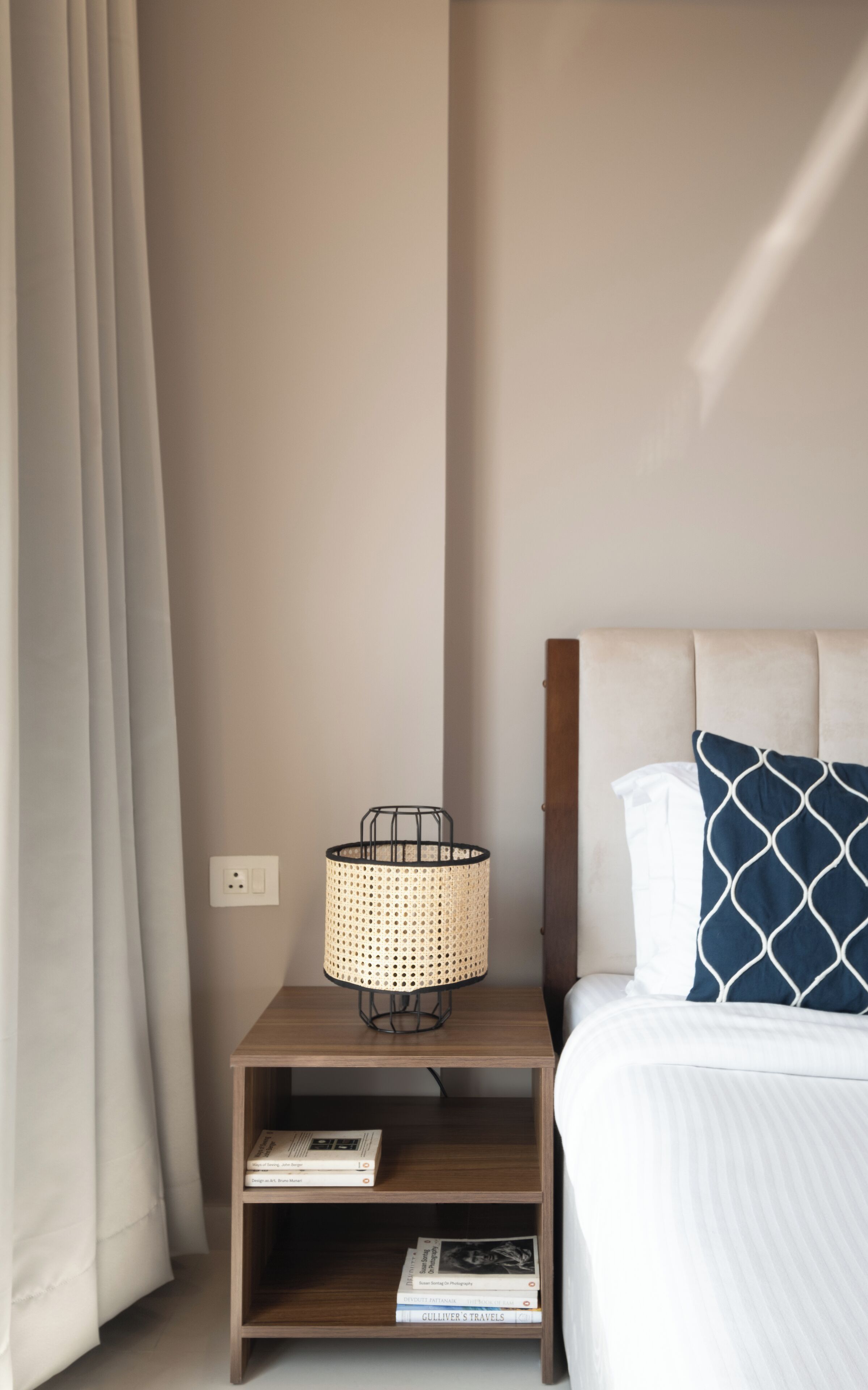 Una cama bien hecha con un cojín estampado, una lámpara de noche en una mesita de madera y una estantería con libros.