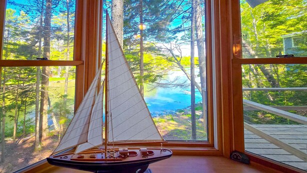 Un modelo de velero dentro de una cabina con ventanas grandes que dan a un bosque y una masa de agua.