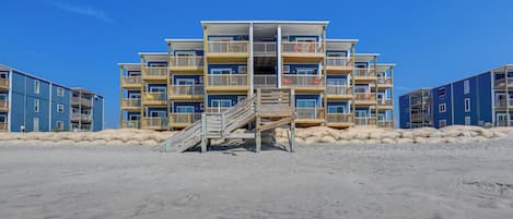 En flervåningsbyggnad som ligger vid stranden, med balkonger, en trappa som leder till entrén och en sandstrand framför.