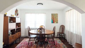 Un comedor con una mesa redonda, sillas de madera, un tapete colorido, un armario de madera y una ventana con cortinas translúcidas.