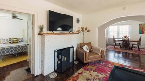 Una sala de estar con chimenea, televisor montado en la pared, sofá, mesa de comedor con sillas y una alfombra colorida.