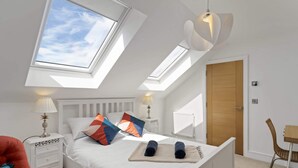 Un dormitorio con tragaluz, una cama con almohadas, una mesita de noche, una silla y una puerta de madera.