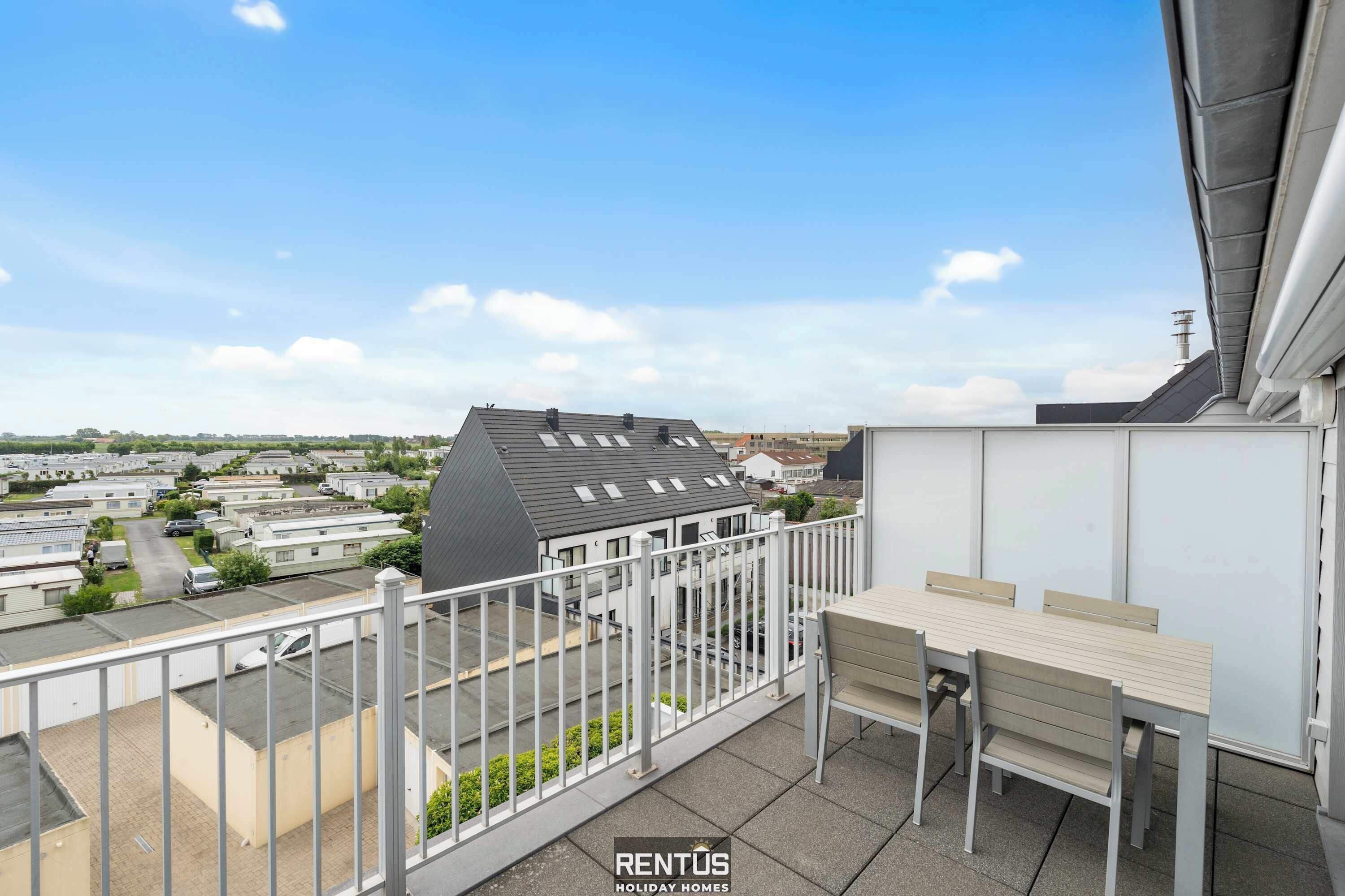 A balcony with a table and chairs, overlooking a residential area with houses and a parking lot.