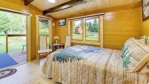 A wooden cabin bedroom with a bed, a chair, a small table, and a window overlooking a yard. - On Hobby Farm: Whidbey Island Tiny Home (Clinton)