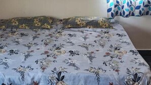 Une pièce avec un lit recouvert d’une couvre-lit à motifs floraux, une fenêtre ornée d’un rideau à motifs de feuilles et une prise électrique au mur.