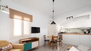 A modern living room with a sofa, a coffee table, and a TV. There is a kitchen area with a dining table and chairs.