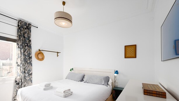A modern bedroom with a large bed, a hanging light fixture, a window with curtains, and a bedside table with a book and a lamp.