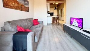 A modern living room with a grey sofa, red cushions, a flat-screen TV, and a kitchen in the background.