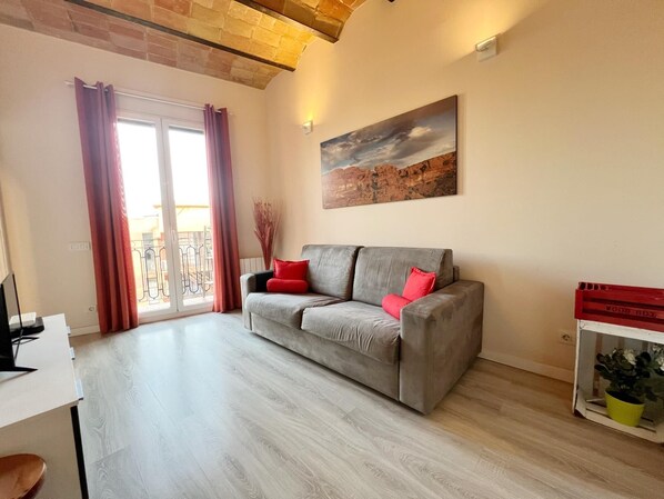 Ein Wohnzimmer mit grauem Sofa, roten Kissen, einem Gemälde, Holzboden und einem Balkon mit roten Vorhängen.