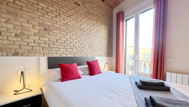 A hotel room with a brick wall, a large bed with white bedding and red pillows, a bedside table with a lamp, and a view of the outside through a window with pink curtains.