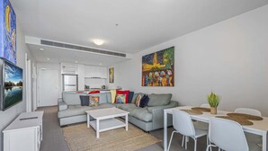 A modern living room with a grey sofa, white coffee table, and a dining area with white chairs and a table. - Docklands Waterfront Elegance with Cityscape Views (Docklands)
