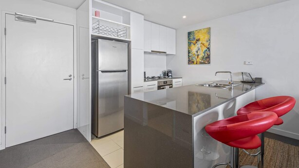A modern kitchen with a stainless steel refrigerator, white cabinets, a central island with bar stools, and a painting on the wall. - Docklands Waterfront Elegance with Cityscape Views (Docklands)