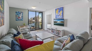A modern living room with a sofa, a coffee table, and a TV. There are paintings on the walls and a balcony with a view of the city. - Docklands Waterfront Elegance with Cityscape Views (Docklands)