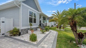 A modern house with a grey exterior, a covered porch, and a landscaped garden with palm trees and shrubs.