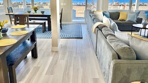 Un salón amplio con un sofá grande, una mesa de comedor con individuales y vistas a la playa a través de grandes ventanales.