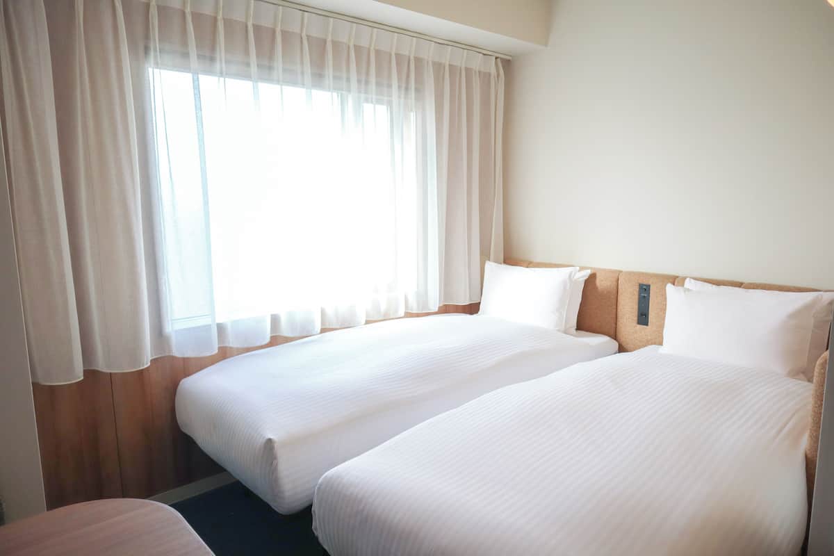 A hotel room with two single beds, a wooden headboard, a window with sheer curtains, and a small bedside table.