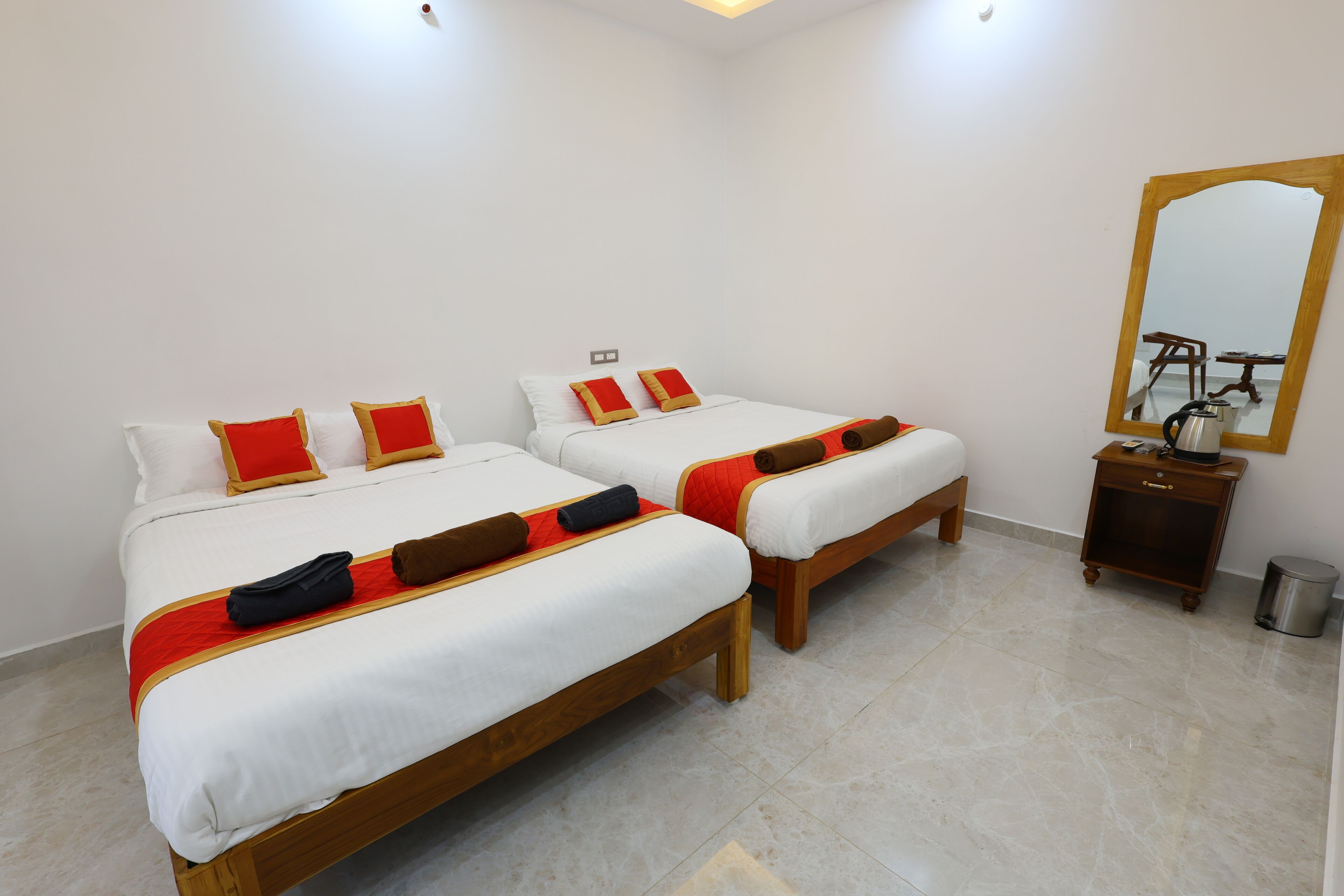 A hotel room with two beds, each with white bedding and red and brown pillows. There is a wooden nightstand with a lamp and a trash bin.