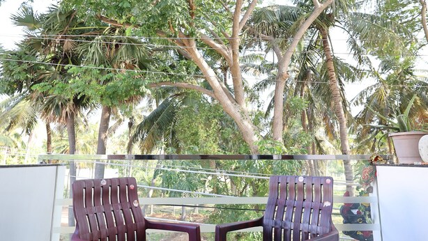 A balcony with two wooden chairs and a glass table, overlooking a lush garden with tall trees and a clear sky. - Star Eden Residency (Chinna Veerampattinam)
