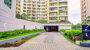 Un edificio moderno de varios pisos con un área de entrada ajardinada, que incluye un sendero pavimentado y follaje.