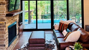 A spacious living room with a stone fireplace, leather sofa, and large windows offering a view of a swimming pool and trees.