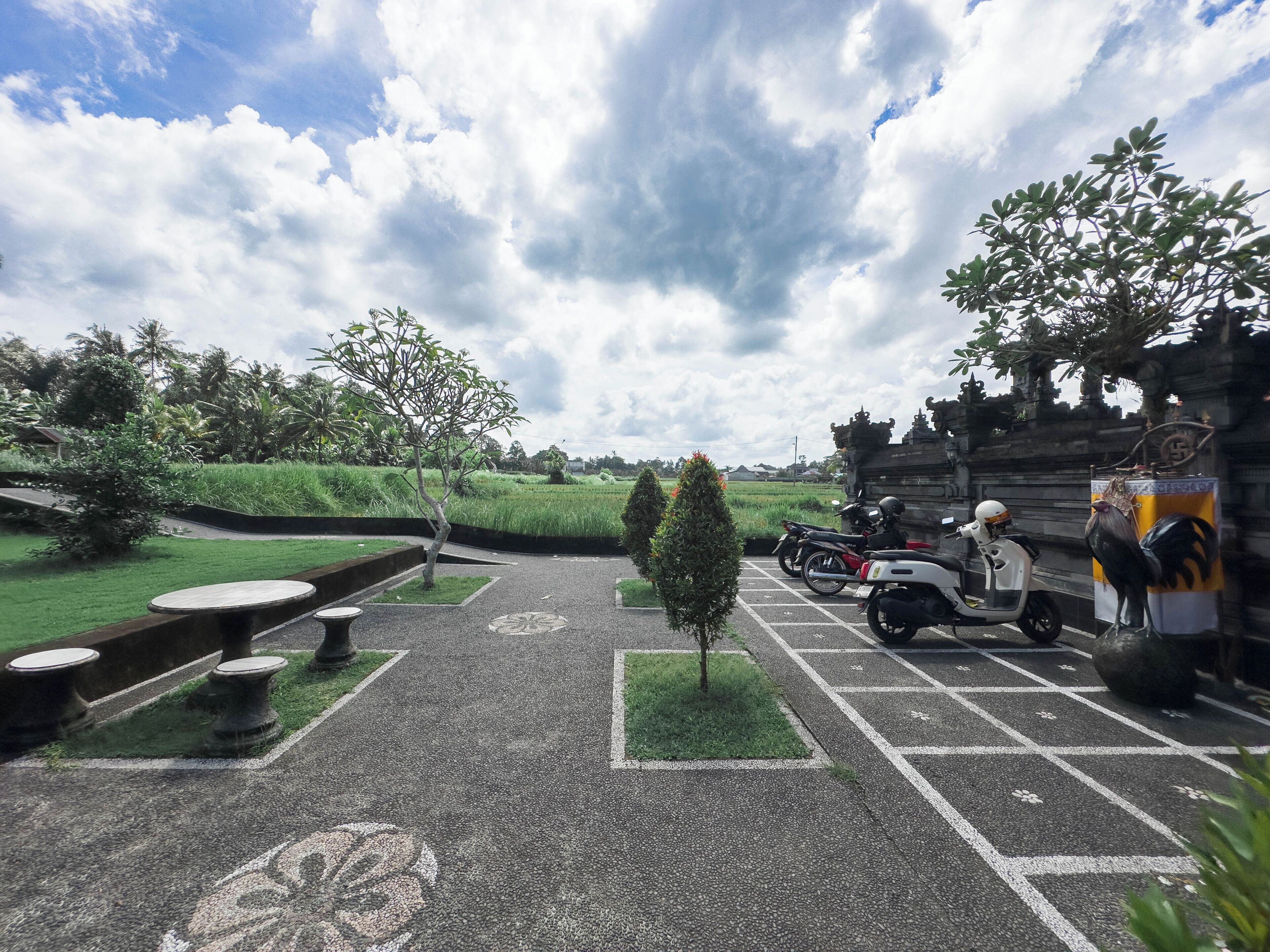 A parking area with scooters, a small tree, and a pathway leading to a building with traditional architecture.