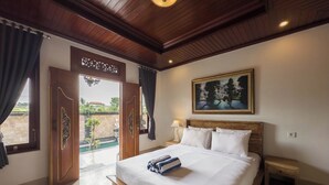 A bedroom with a wooden bed, a nightstand, a painting, and a view of a garden through a door. - Tiara Guest House Ubud (Ubud)